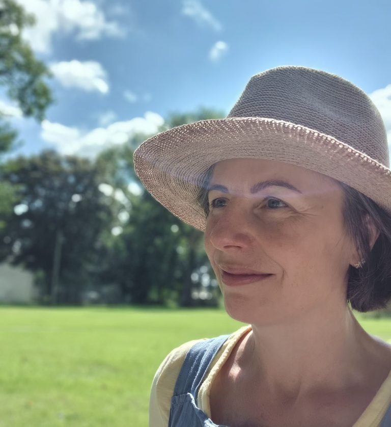 a photo of Kirstie (portrait) in a sun hat outside on a sunny day with the summer sky and lush trees in the background. 
